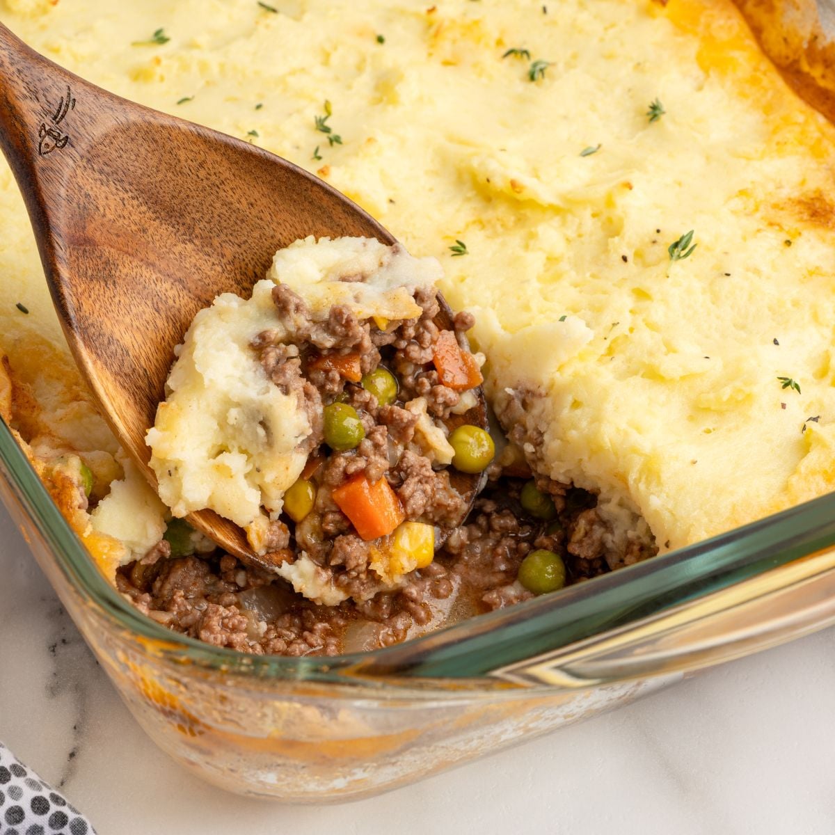 A wooden spoon lifts a serving of Shepherd's Pie from a glass baking dish, revealing layers of mashed potatoes, ground meat, peas, carrots, and corn—classic comfort in every bite and perfect for anyone searching for the best shepherd's pie recipe.