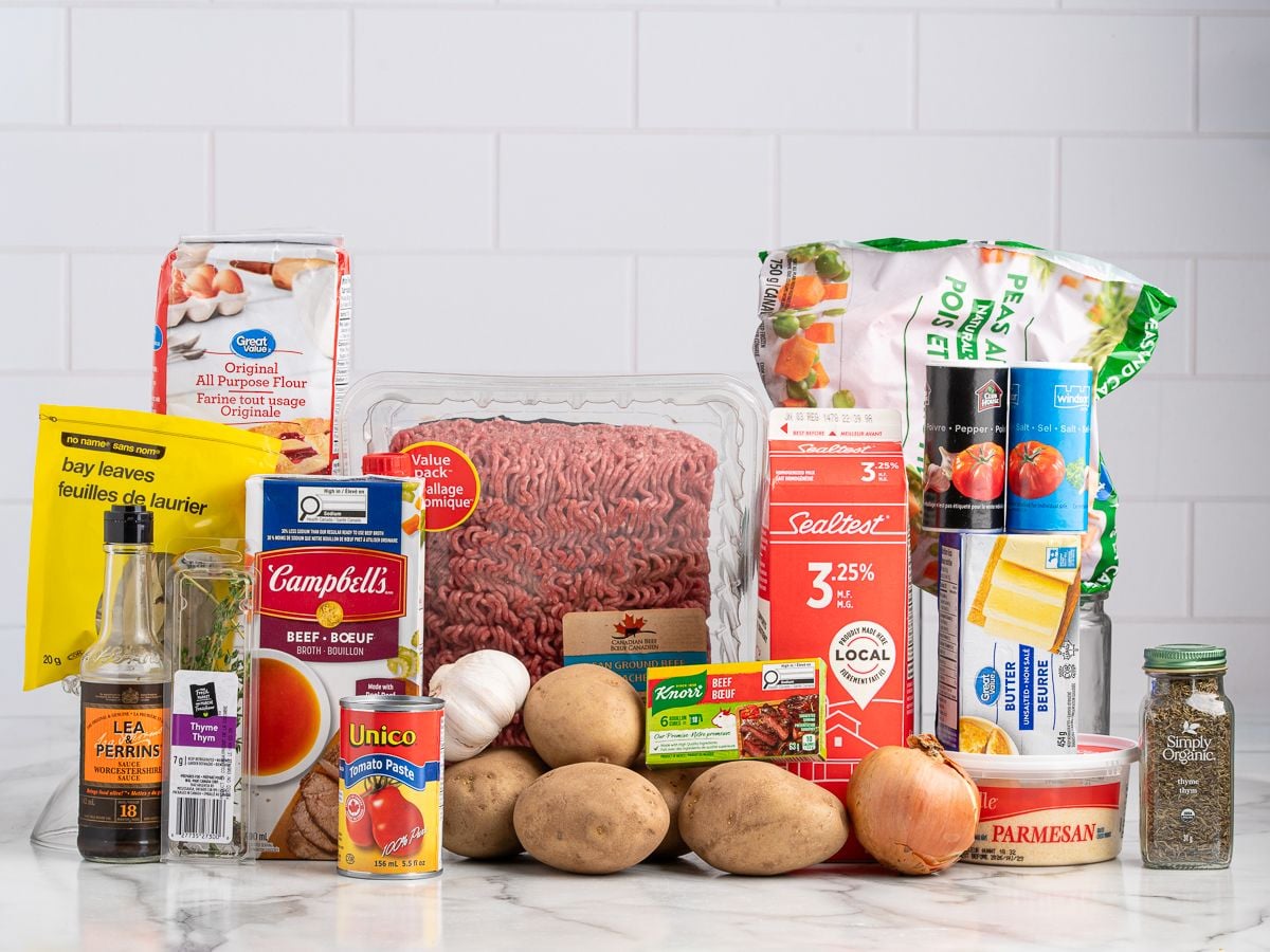 A variety of grocery items on a counter, perfect for making the best shepherd's pie recipe—ground beef, potatoes, flour, canned tomatoes, frozen peas and carrots, onion, garlic, cheese, butter, milk, bay leaves, beef broth, Worcestershire sauce, and seasonings.