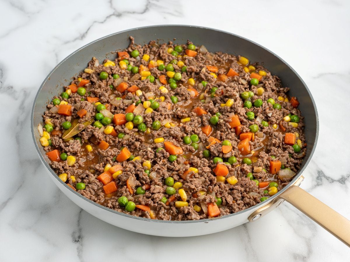 A skillet filled with cooked ground beef mixed with peas, carrots, corn, and chopped onions sits on a white marble countertop—the perfect base for the best shepherd's pie recipe.
