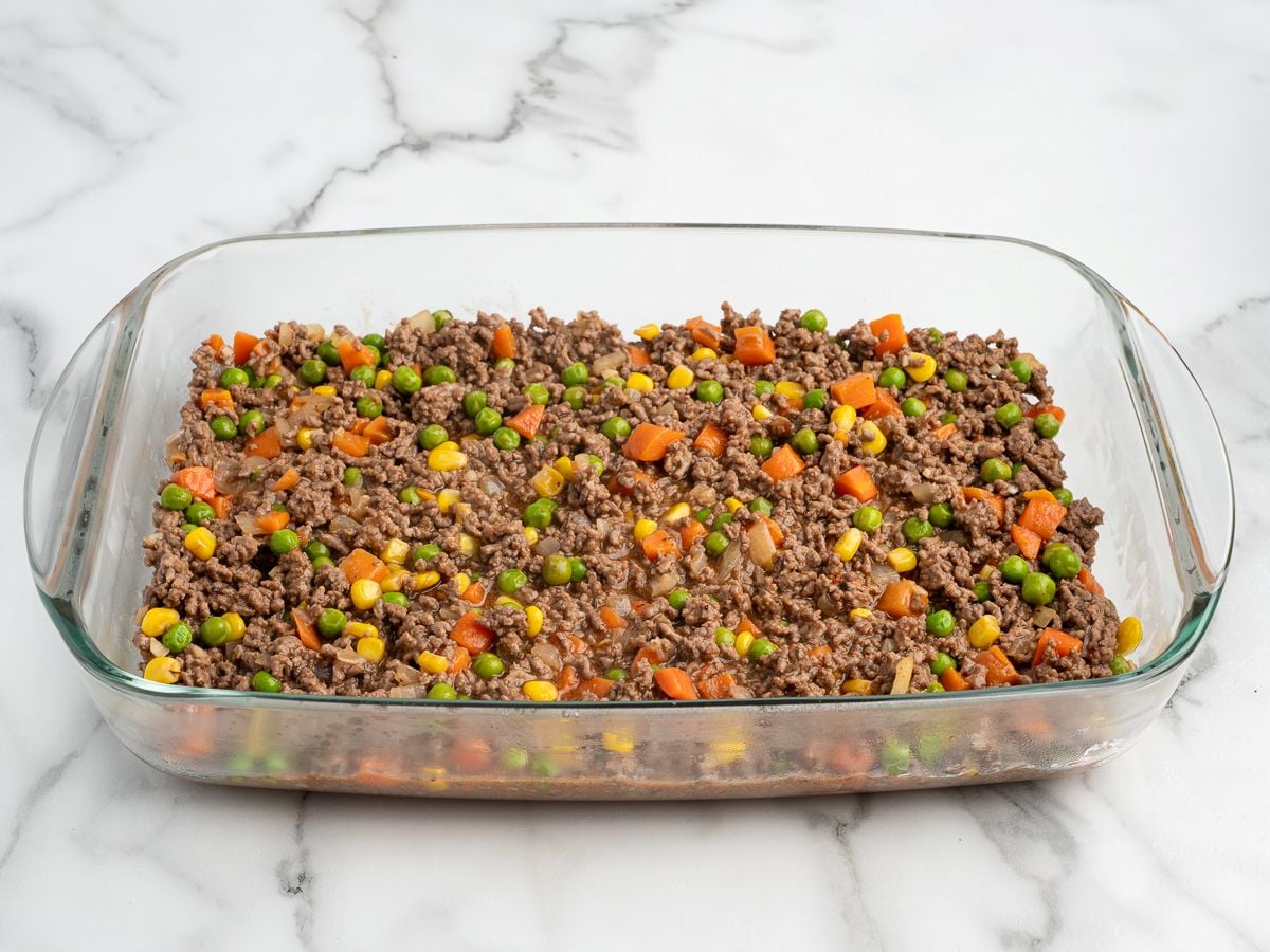 A glass baking dish filled with a savory shepherd's pie recipe—ground beef, peas, carrots, corn, and onions—sits on a white marble surface.