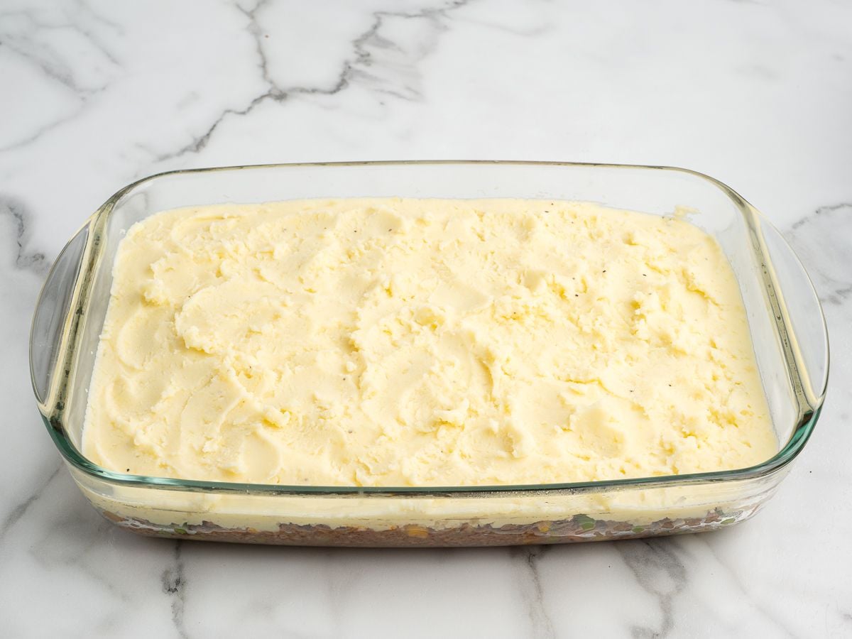 A glass baking dish filled with shepherd’s pie, made from the best shepherd's pie recipe and topped with a smooth layer of mashed potatoes, sits on a white marble surface.