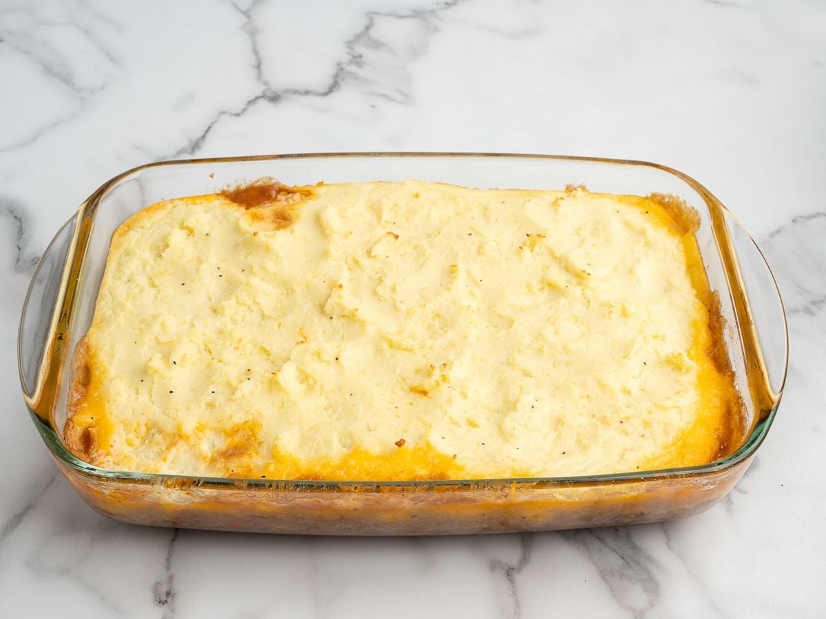 A glass baking dish filled with a freshly baked shepherd's pie, topped with a smooth, golden layer of mashed potatoes, on a white marble surface—just like the best shepherd's pie recipe.
