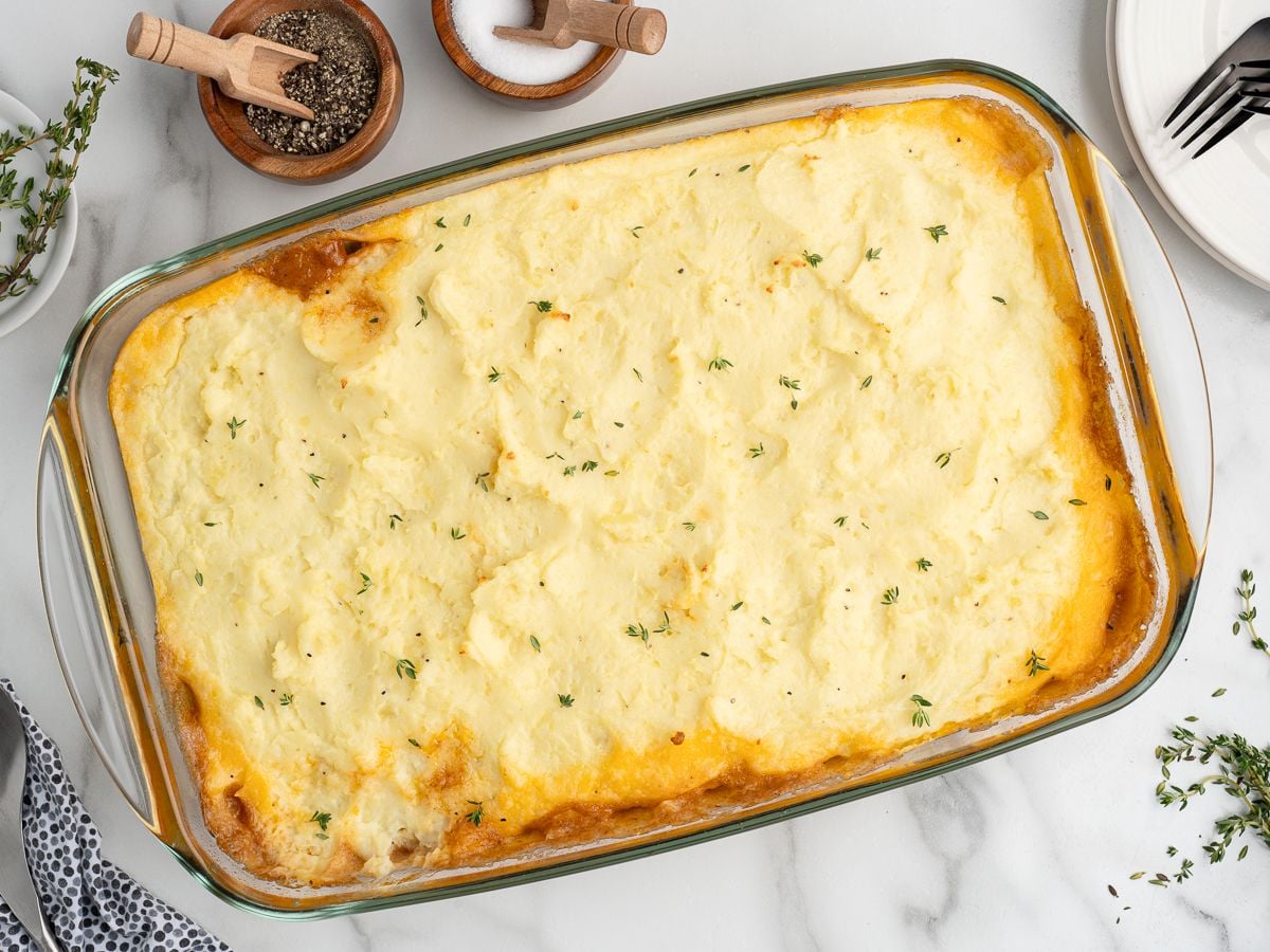 A glass baking dish filled with the best shepherd's pie recipe, topped with mashed potatoes and fresh thyme. Small bowls of salt and pepper with wooden spoons sit nearby on a marble surface.