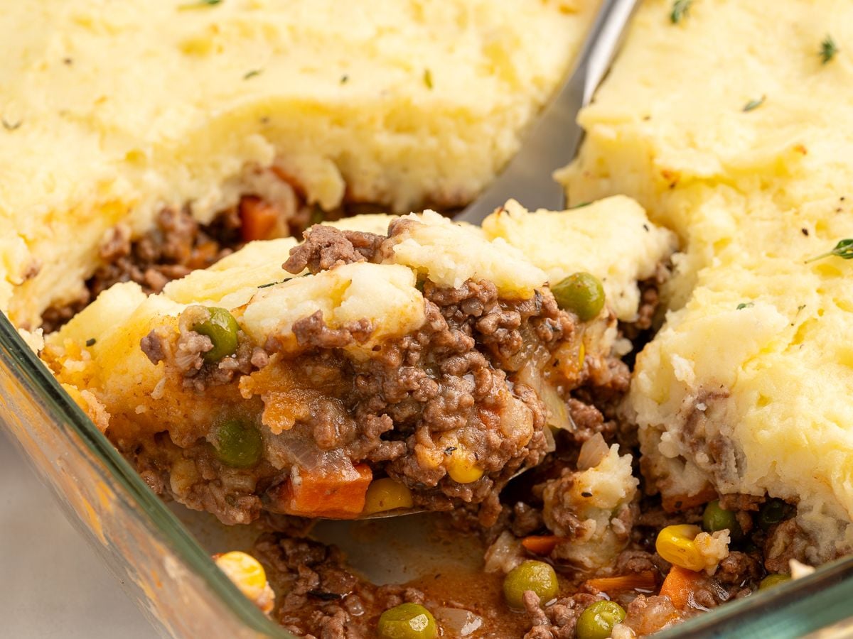 A close-up of a serving of shepherds pie being lifted from a glass baking dish, showing the classic shepherd's pie recipe with seasoned ground beef, peas, carrots, and corn beneath a layer of creamy mashed potatoes.