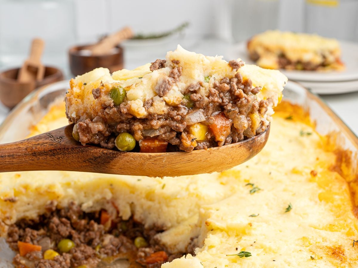 A wooden spoon lifts a serving of the best Shepherd's Pie recipe from a glass baking dish, revealing layers of mashed potatoes, ground meat, peas, carrots, and corn, with more Shepherd's Pie visible in the background.