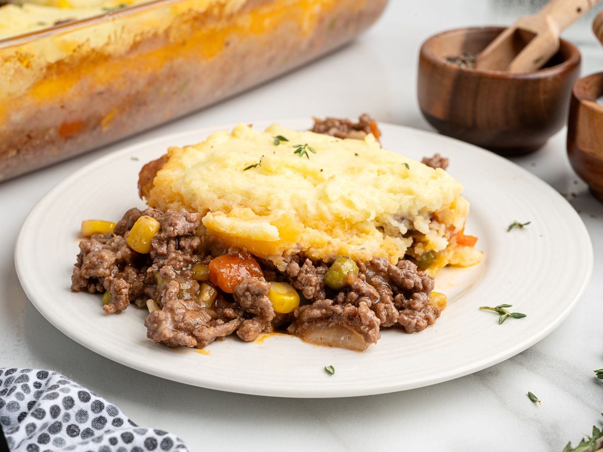 A serving of shepherd’s pie on a white plate, showing layers of ground meat, mixed vegetables like corn and carrots, and creamy mashed potatoes. The classic shepherd’s pie recipe sits beside wooden spice containers in the background.
