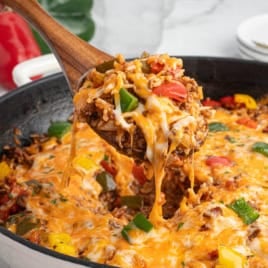 A wooden spoon lifts a cheesy serving of Stuffed Pepper Casserole with rice, ground meat, bell peppers, and melted cheese from a skillet.