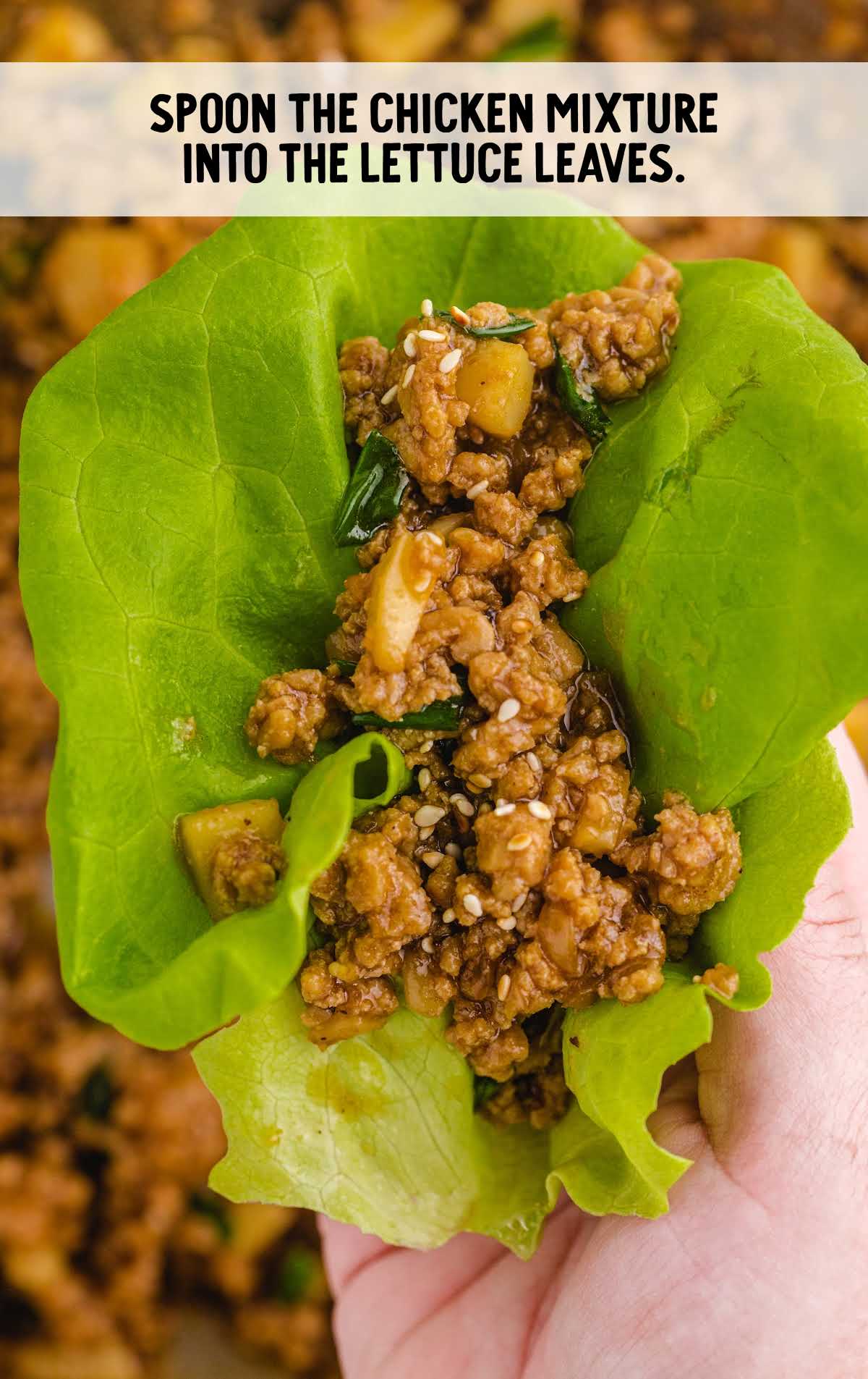 Close-up of hand holding a single lettuce wrap filled with chicken mixture, sesame seeds, and diced water chestnuts.