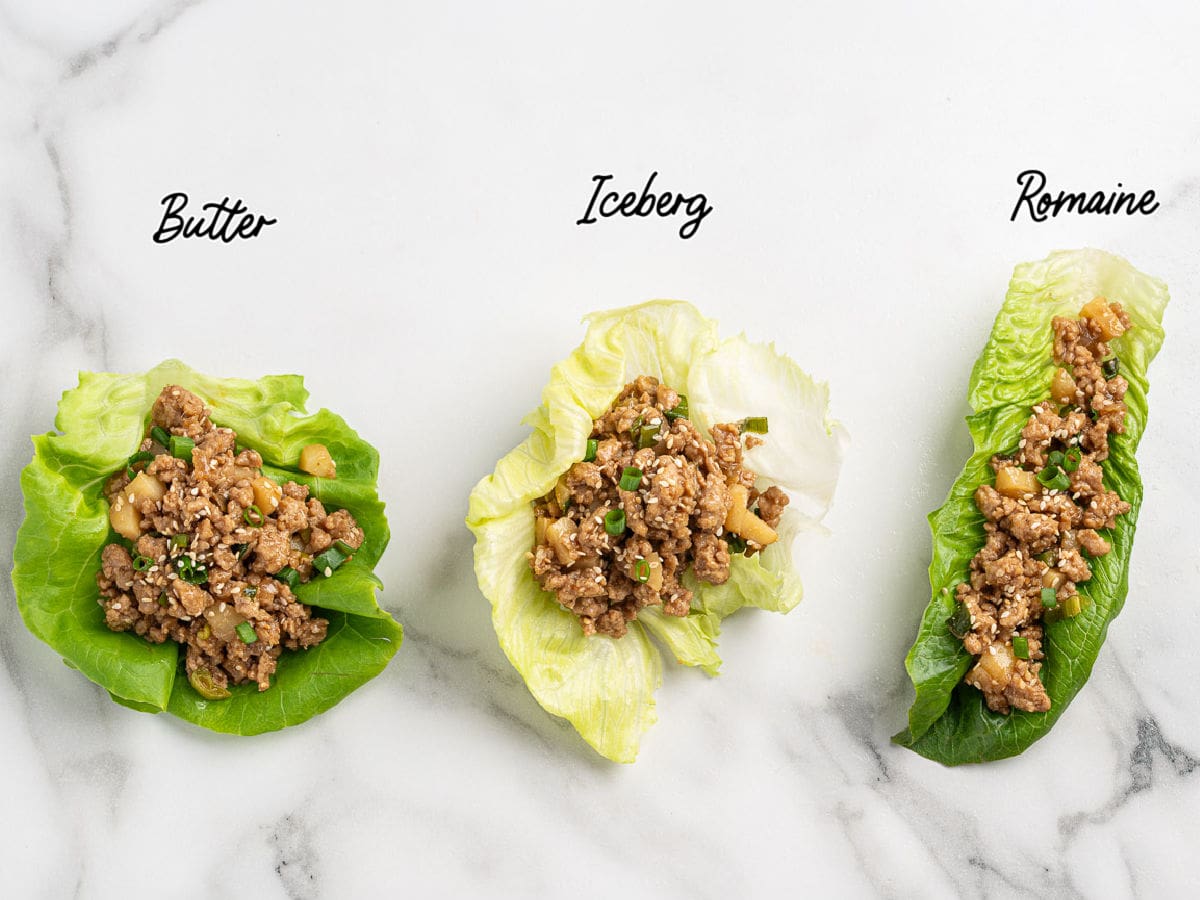 Three lettuce wraps, inspired by a classic Asian Chicken Lettuce Wrap Recipe, are displayed side by side on a marble surface. Each wrap is filled with seasoned ground meat and uses butter, iceberg, or romaine lettuce, with each variety labeled above the corresponding Chicken Lettuce Wrap.