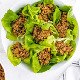 White bowl filled with Asian chicken lettuce wraps, ready to serve, on a white marble background.