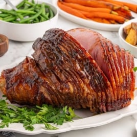 Fully glazed spiral ham on a white platter with carrots, green beans, and potatoes in the background.