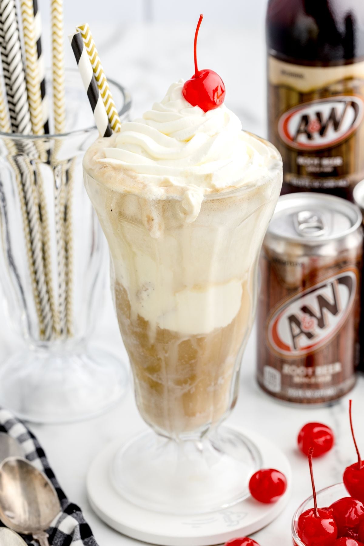 A Root Beer Float in a tall glass topped with whipped cream and a cherry, with a striped straw. A&W Root Beer cans and bottles, striped straws in a glass, and maraschino cherries are in the background.