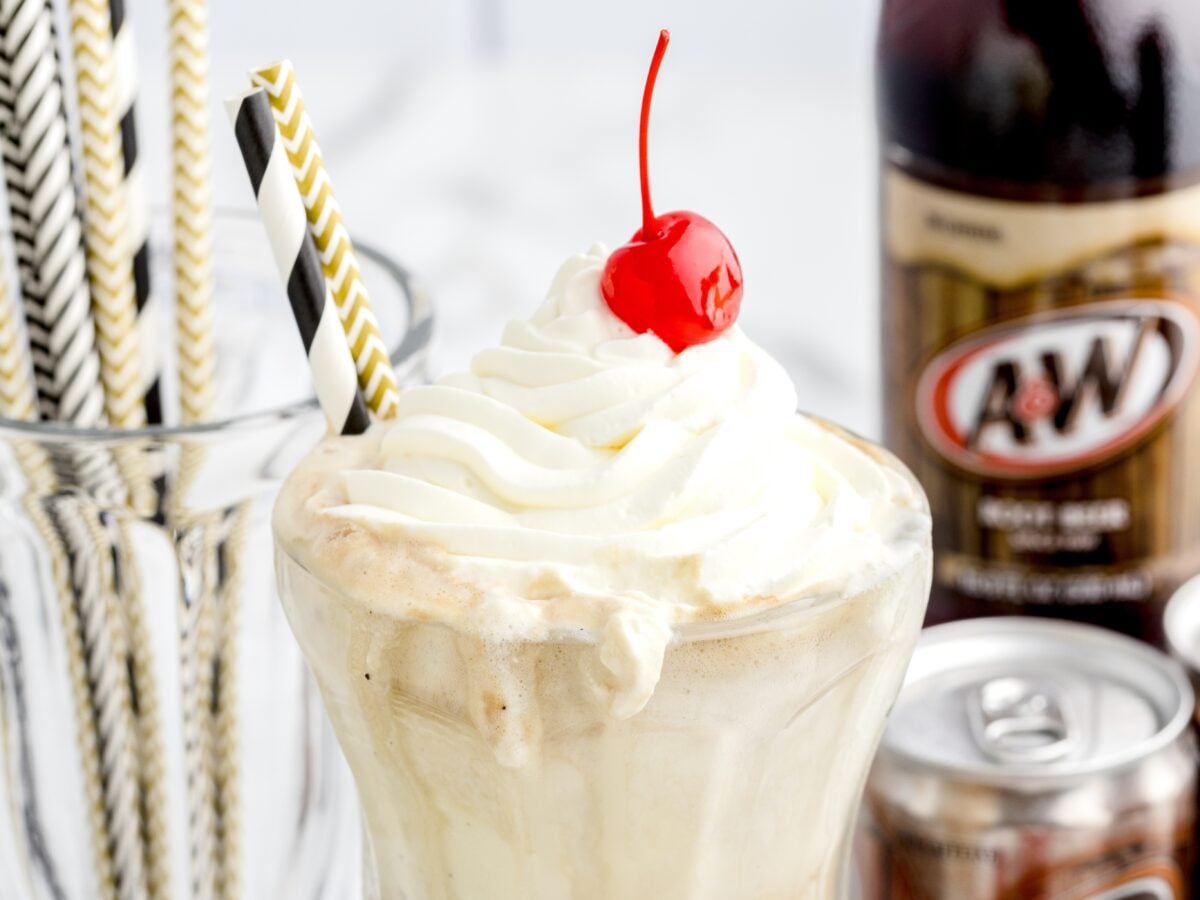 A Root Beer Float in a glass topped with whipped cream and a cherry, with a black and gold striped straw. A&W root beer bottles and cans, plus a jar of straws, are visible in the background.