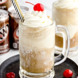 A glass mug of Root Beer Float topped with whipped cream and a cherry, served with a striped straw. Cans of A&W root beer and another float are in the background; a cherry sits on the plate beside the mug.