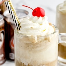 A glass mug filled with a classic Root Beer Float topped with whipped cream and a cherry, complete with two striped straws. Cans of A&W root beer and another frosty mug appear in the background.