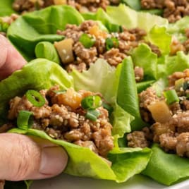 A hand holds a lettuce wrap filled with seasoned ground meat, diced vegetables, green onions, and sesame seedsโjust like what youโd find in a classic Asian Chicken Lettuce Wrap Recipe. Several more chicken lettuce wraps are arranged on a plate in the background.