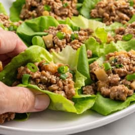 A hand holding a lettuce wrap filled with seasoned ground meat, diced vegetables, green onions, and sesame seeds; more Chicken Lettuce Wraps are arranged on a white plate. Perfect for anyone craving an Asian Chicken Lettuce Wrap Recipe that’s both flavorful and fresh.