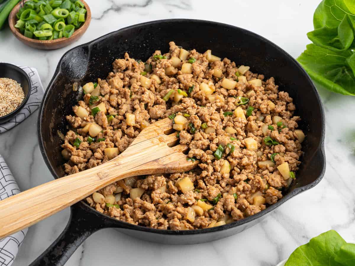 A skillet filled with cooked ground meat, diced vegetables, and herbs, perfect for an Asian Chicken Lettuce Wrap Recipe, with a wooden spatula resting inside. Chopped green onions and lettuce leaves are nearby on the marble countertop.