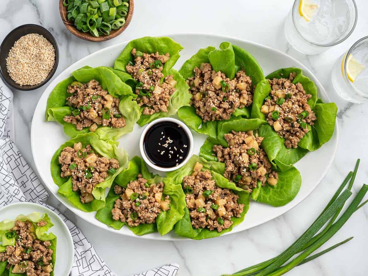 A white platter with Chicken Lettuce Wraps filled with ground meat and diced vegetables, garnished with sesame seeds and chives, surrounds a small dish of soy sauce. Green onions, sesame seeds, and lemon water are beside the platter in this tempting Asian Chicken Lettuce Wrap Recipe.