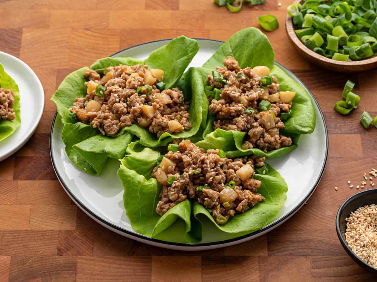 Four lettuce wraps filled with seasoned ground meat and diced vegetables are arranged on a white plate. Reminiscent of a classic Asian Chicken Lettuce Wrap Recipe, these wraps are garnished with chopped green onions and sesame seeds, with extra toppings in small bowls nearby.
