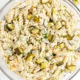 Overhead shot of dill pickle pasta salad in a glass bowl with creamy dressing, chopped pickles, and fresh dill on a white countertop.