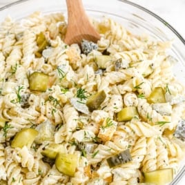 Close-up of dill pickle pasta salad with rotini, chopped pickles, cheddar cheese cubes, and creamy dressing, served in a white bowl with a wooden spoon.