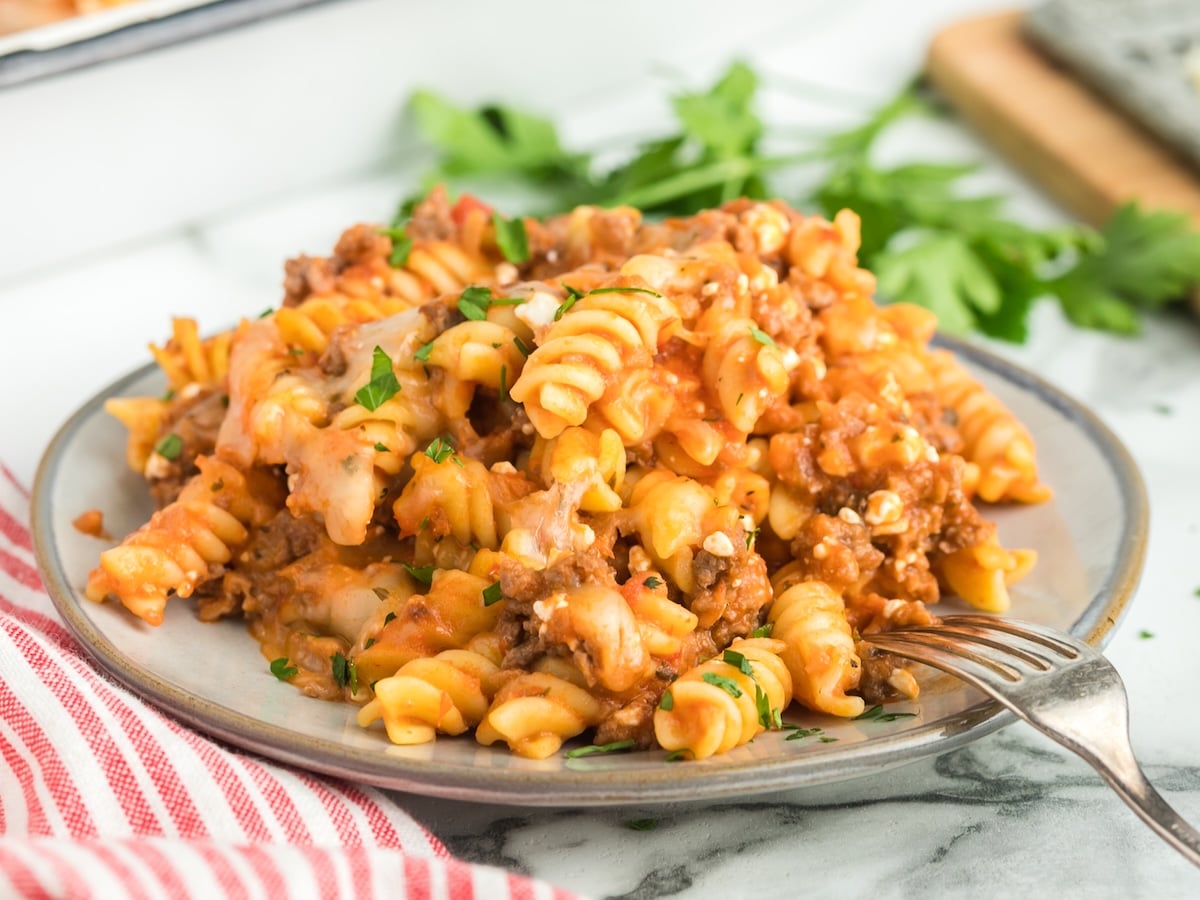 A plate of baked rotini pasta with ground meat, tomato sauce, melted cheese, and parsley garnish&mdash;this hearty Lasagna Casserole sits ready to enjoy. A fork rests on the plate, with a red and white striped towel and fresh herbs in the background.