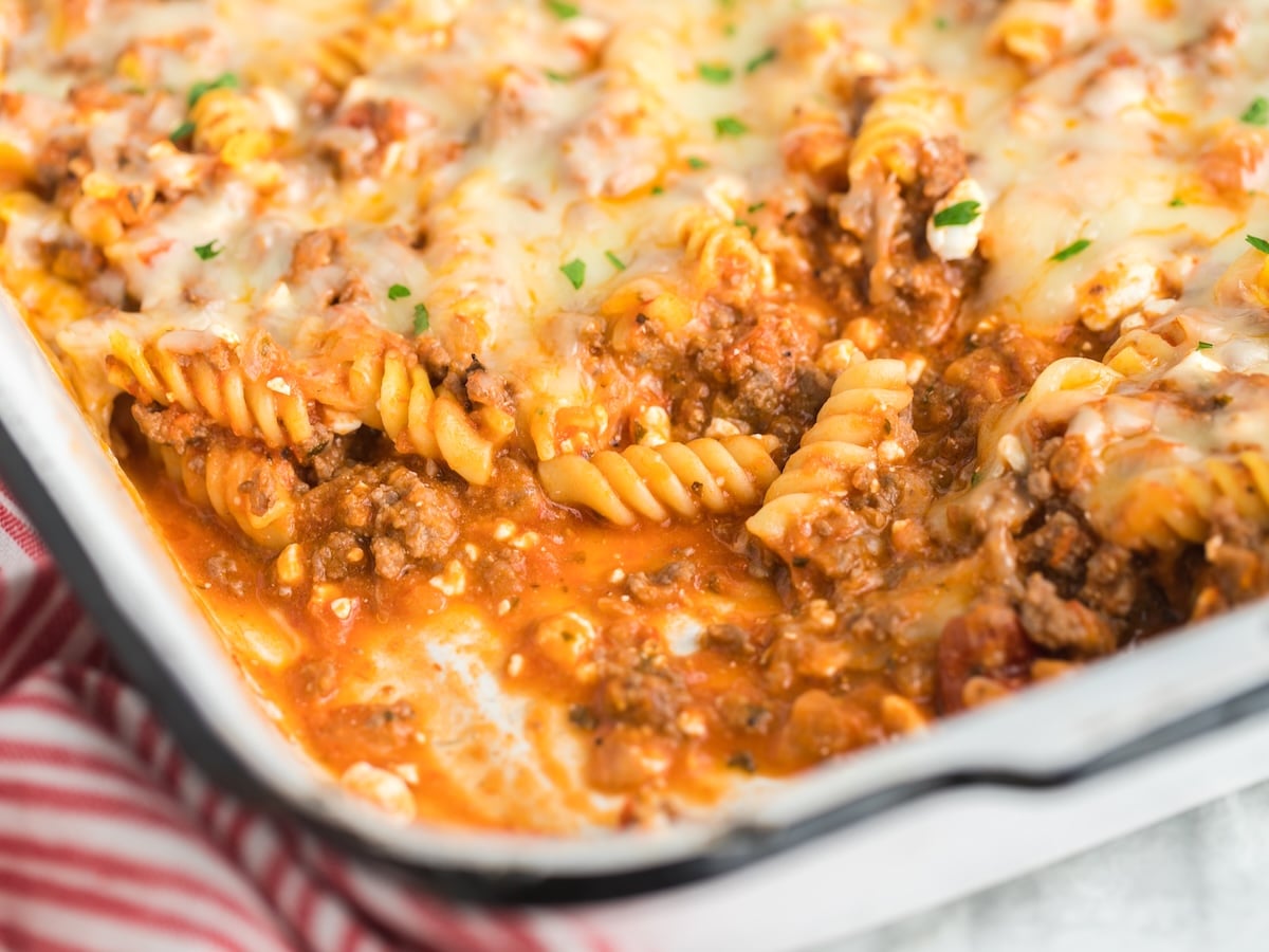 A close-up of a hearty Lasagna Casserole with rotini noodles, ground beef, tomato sauce, and melted cheese in a white baking dish, some portions already served. A red-striped towel is partially visible nearby.