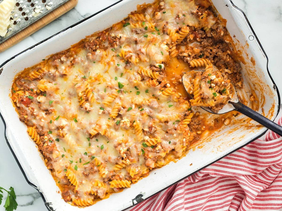 A baked Lasagna Casserole with rotini, ground meat, tomato sauce, and melted cheese sits in a white dish. A serving has been scooped out with a spoon, and a red striped towel is nearby.