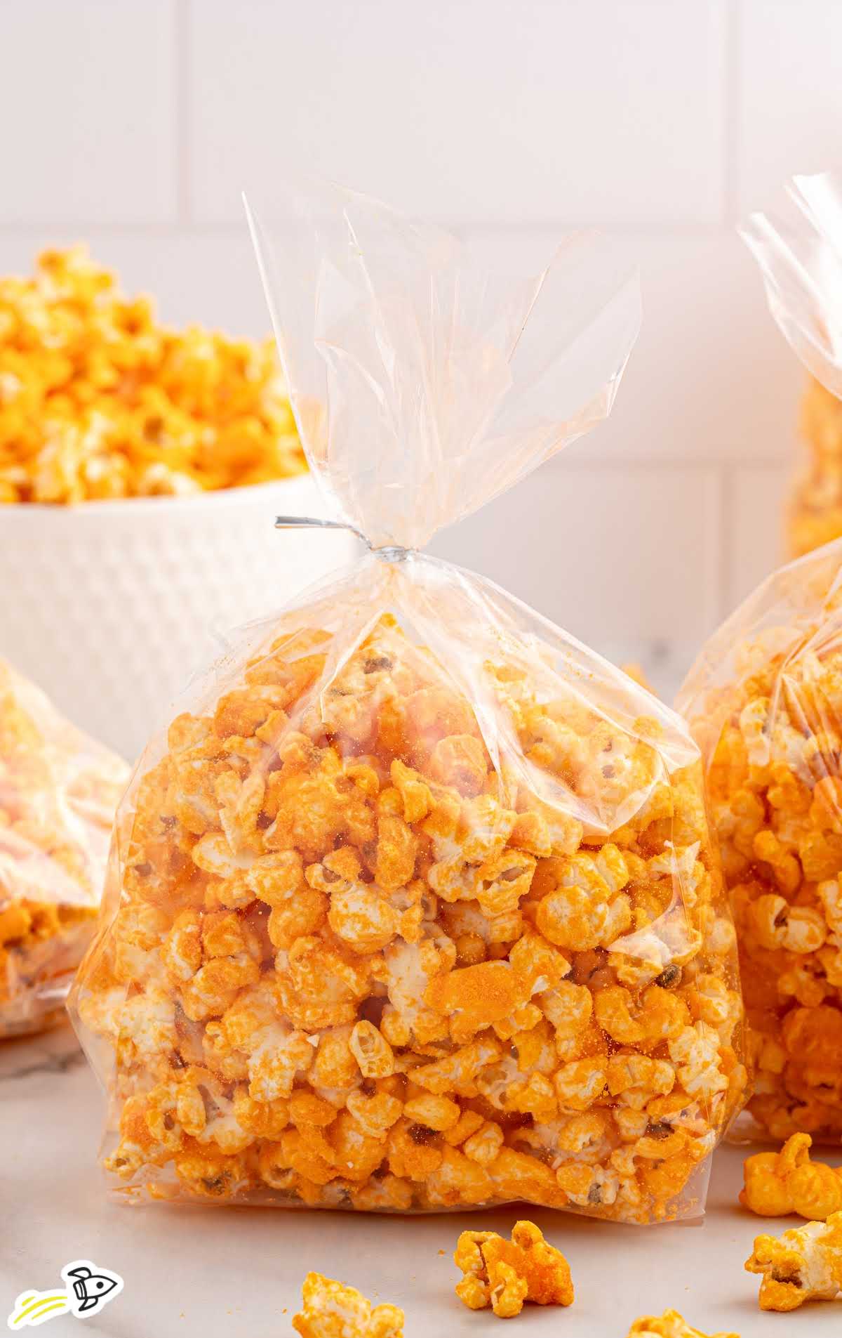 A clear plastic bag filled with homemade cheese popcorn, tied with a silver twist tie, set against a blurred background of more popcorn.