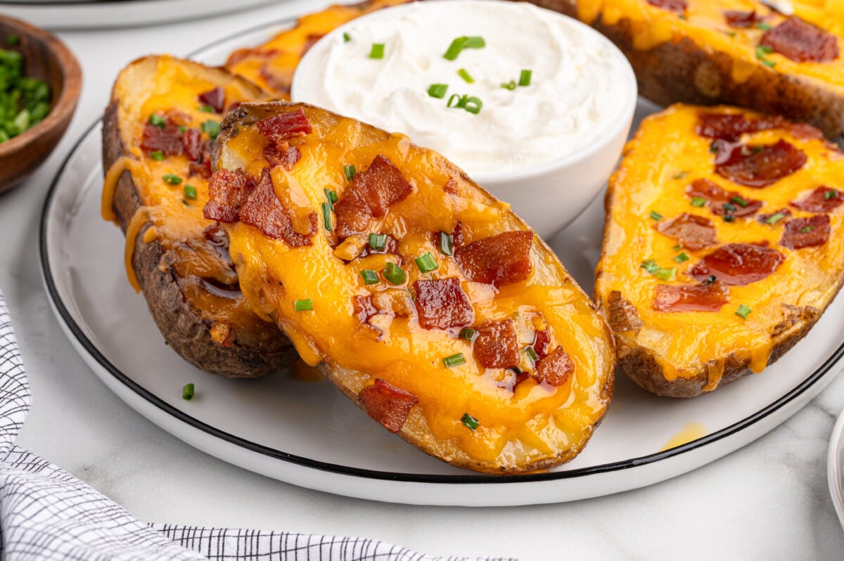 Three baked potato skins topped with melted cheddar cheese, bacon pieces, and chopped chives are arranged on a plate with a bowl of sour cream for dipping in the center.