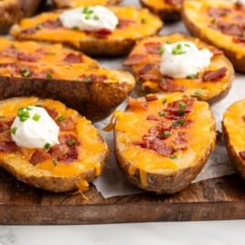 Baked Potato Skins topped with melted cheddar cheese, bacon pieces, sour cream, and chopped chives, arranged on a wooden board lined with parchment paper for a savory appetizer everyone will love.