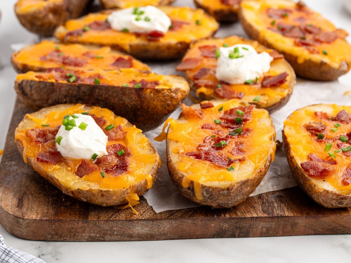 Baked Potato Skins topped with melted cheddar cheese, bacon pieces, sour cream, and chopped chives, arranged on a wooden board lined with parchment paper for a savory appetizer everyone will love.