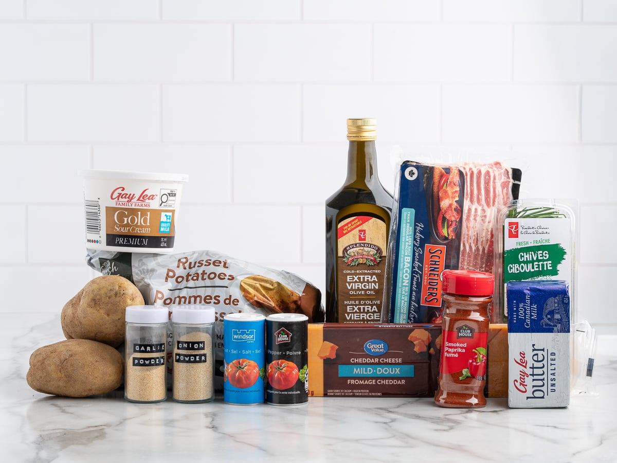 A group of ingredients for delicious Baked Potato Skins sits on a marble counter, including russet potatoes, sour cream, extra virgin olive oil, bacon, butter, cheddar cheese, canned tomatoes, dried chives, and assorted spices in small jars.