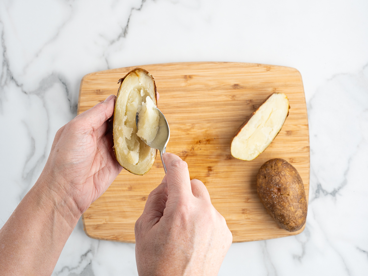A person scoops out the inside of a baked potato with a spoon over a wooden cutting board, preparing baked potato skins. The board also holds another halved potato and a whole potato.