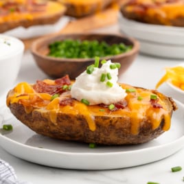 Loaded Baked Potato Skins topped with melted cheddar cheese, bacon pieces, a dollop of sour cream, and chopped chives, served on a white plate with bowls of sour cream, chives, and shredded cheese in the background.