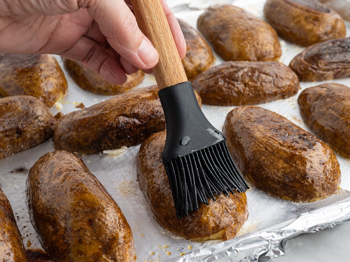 A hand uses a black silicone brush to coat Baked Potato Skins with oil or butter on a foil-lined baking sheet.