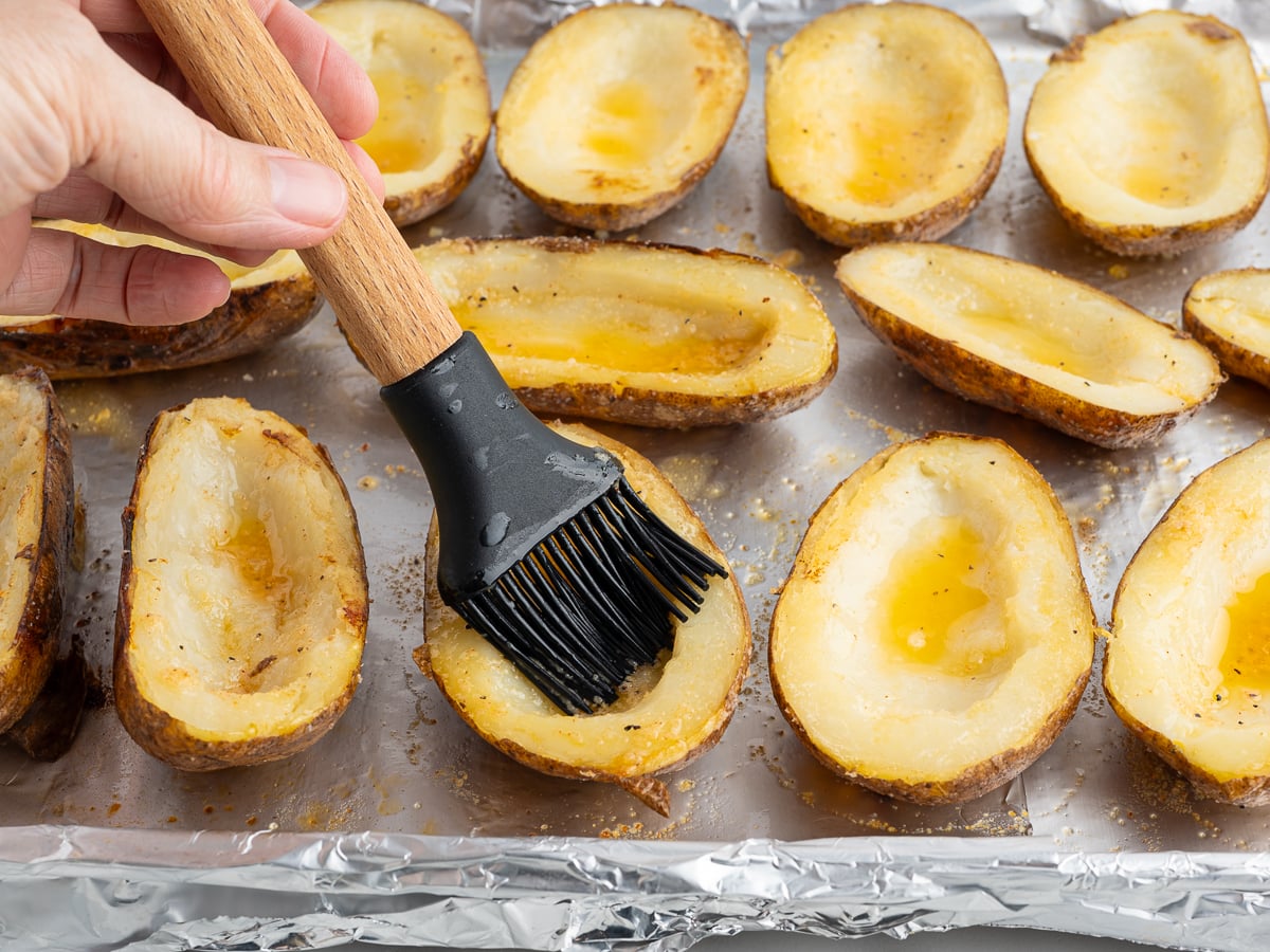 A hand uses a basting brush to coat Baked Potato Skins with oil on a foil-lined baking sheet. The potato skins are arranged cut-side up, looking seasoned and ready for further cooking.