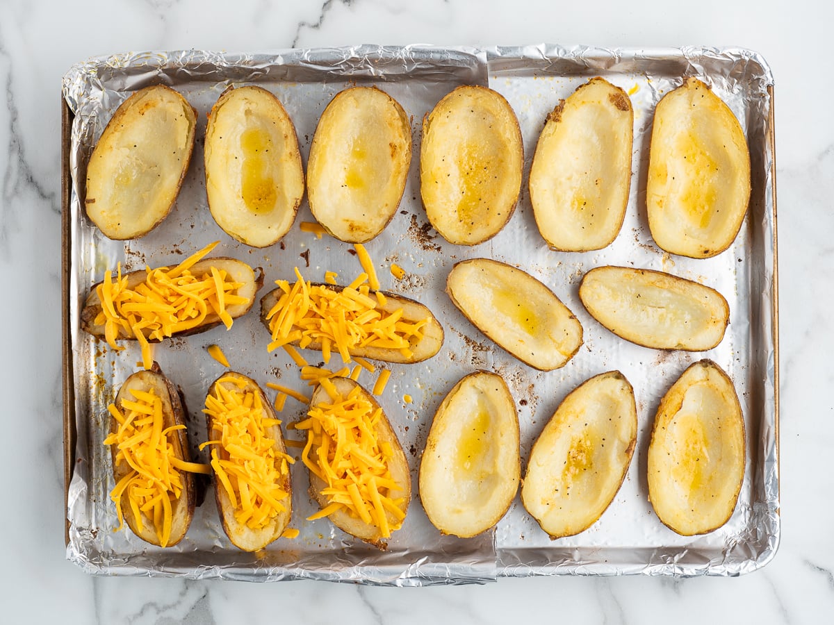A baking sheet lined with foil holds halved baked potatoes; some are plain, while others are filled with shredded cheddar cheese—delicious Baked Potato Skins ready for further cooking.