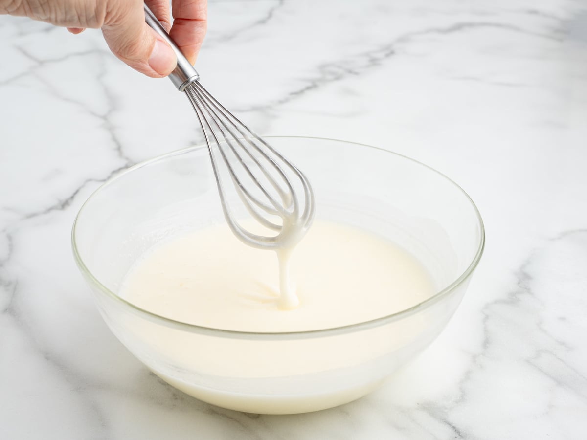 A hand holds a whisk above a clear glass bowl filled with smooth, white Lemon Drop Cakes mixture on a marble countertop.