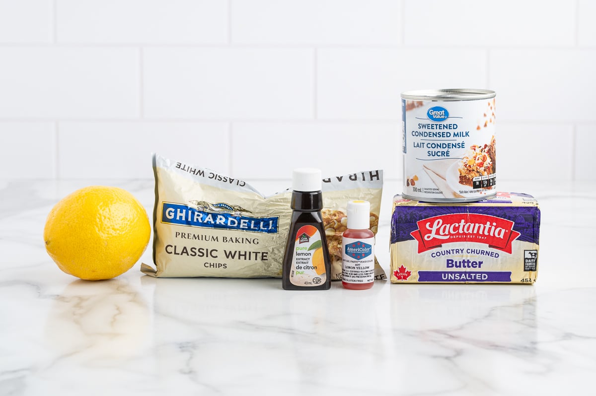 A lemon, white chocolate chips, vanilla extract, red food coloring, sweetened condensed milk, and a stick of unsalted butter—all the essentials for a delicious Lemon Fudge Recipe—are arranged on a marble countertop with a white tile background.
