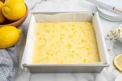 A square baking pan lined with parchment paper is filled with unbaked lemon fudge recipe batter, sprinkled with lemon zest. Fresh lemons, a bowl, a knife, plates, and a checkered cloth are nearby on a marble countertop.