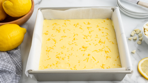 A square baking pan lined with parchment paper is filled with unbaked lemon fudge recipe batter, sprinkled with lemon zest. Fresh lemons, a bowl, a knife, plates, and a checkered cloth are nearby on a marble countertop.