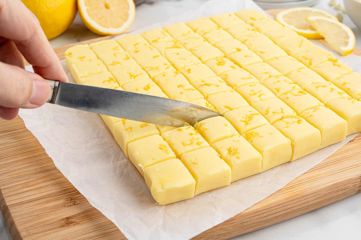 A hand uses a knife to cut lemon fudge into small squares on parchment paper, with lemon halves and zest in the background, showcasing the freshness of this Lemon Fudge Recipe.