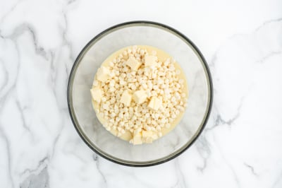 A glass bowl filled with white chocolate chips, small cubes of butter, and a creamy liquid, perfect for starting a delicious Lemon Fudge Recipe, sits on a white marble surface.