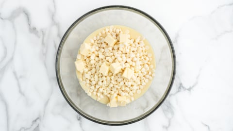A glass bowl filled with white chocolate chips, small cubes of butter, and a creamy liquid, perfect for starting a delicious Lemon Fudge Recipe, sits on a white marble surface.
