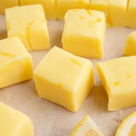 Yellow lemon fudge squares are arranged on parchment paper, with small lemon zest pieces on top. A lemon slice is partially visible in the bottom right corner, highlighting this delicious Lemon Fudge Recipe.