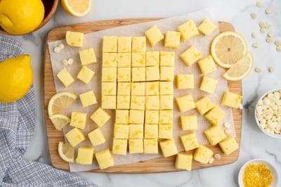 A wooden board topped with neatly cut yellow Lemon Fudge Recipe squares, surrounded by lemon slices, lemon zest, white chocolate chips, and whole lemons on a marble surface.
