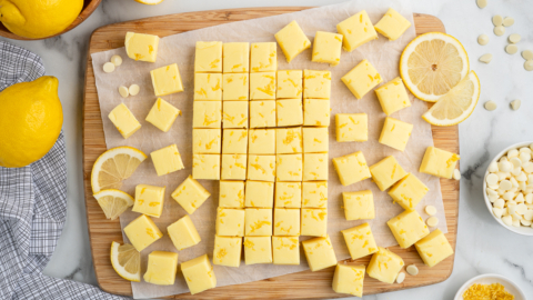 A wooden board topped with neatly cut yellow Lemon Fudge Recipe squares, surrounded by lemon slices, lemon zest, white chocolate chips, and whole lemons on a marble surface.