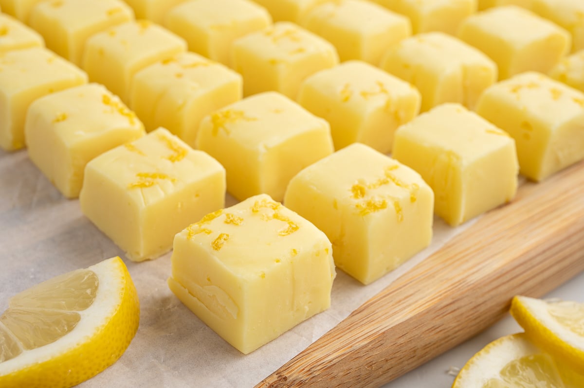 Rows of creamy yellow lemon fudge squares, perfected by this Lemon Fudge Recipe, are topped with lemon zest and arranged neatly on parchment paper, with fresh lemon wedges placed nearby.